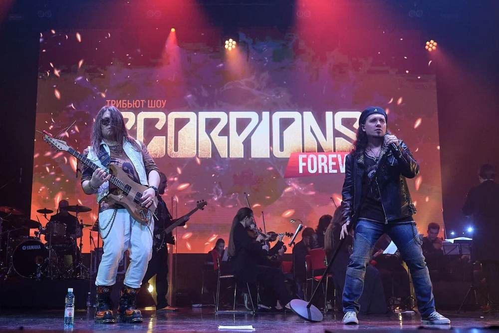 Гитарист и вокалист Rock Believers на сцене, за ними симфонический оркестр